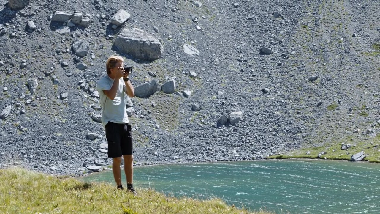 Un giovane in abiti da trekking si ferma su una sponda erbosa per fotografare un lago di montagna ventoso contro un ripido pendio detritico