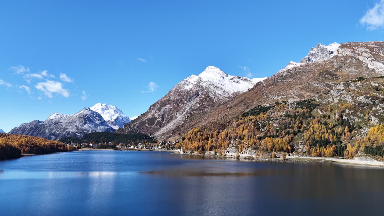 A wide, low aerial view of Lake Silvaplana in the Upper Engadin, Schweiz. The deep blue water reflects a brilliant sky, framed by golden larch slopes and majestic snow-capped mountains