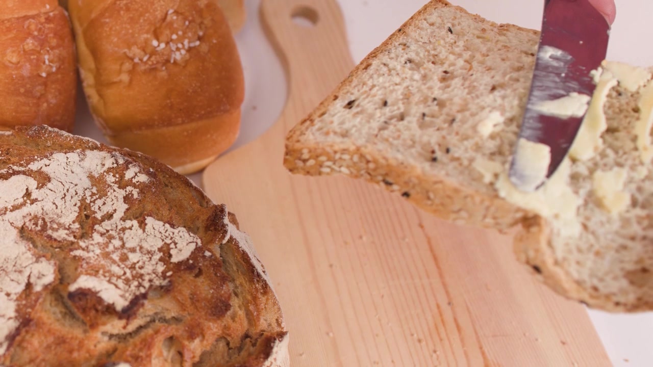 Close-up shot of a hand spreading peanut butter on multigrain bread with assorted bakery loaves.