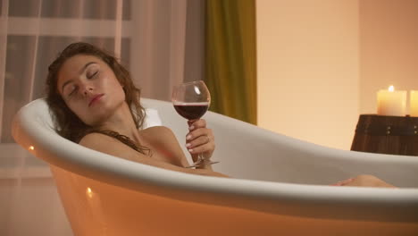 a young beautiful caucasian brunette lies in the bathroom by candlelight in a pleasant evening atmosphere resting from stress and relaxing drinking red wine from a glass