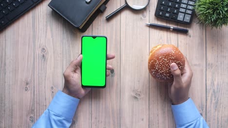 persona sosteniendo un teléfono inteligente con una pantalla verde y una hamburguesa en un escritorio de madera