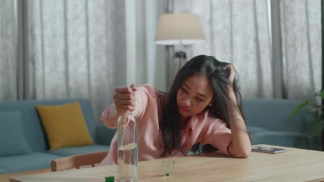 술에 취한 우울한 아시아 여성이 테이블 위에 스마트폰을 들고 집에 있는 병에서 보드카를 마시고 있습니다.