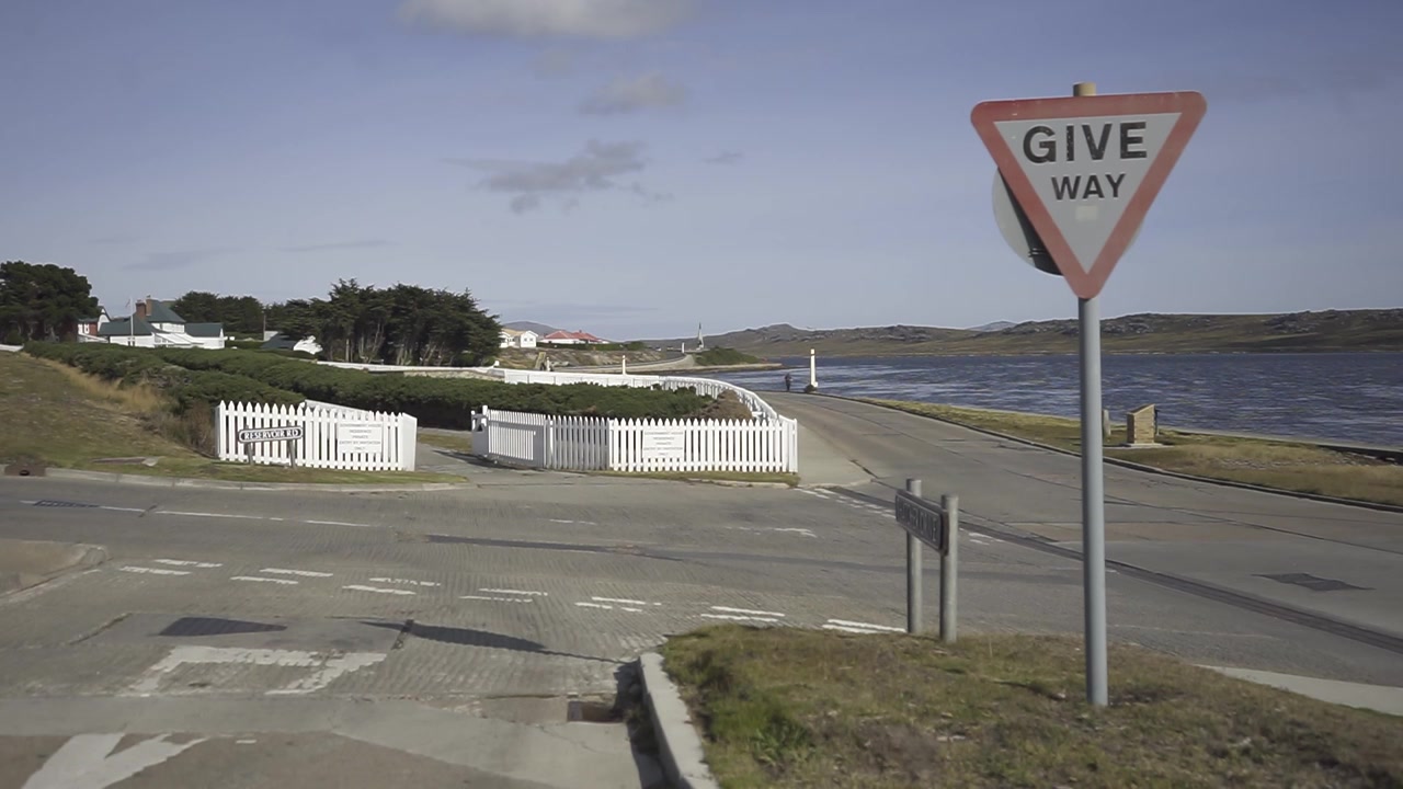 Windböen an einem Vorfahrt-achten-Schild an einer ländlichen Kreuzung nahe dem Meer, umgeben von Gras und Bäumen