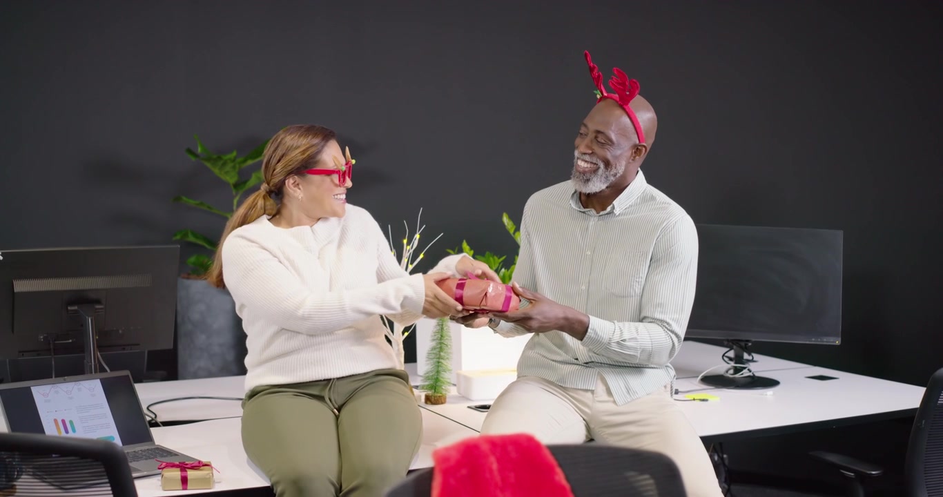 African American coworkers in antlers swapping gifts at desk man offering woman taking, celebrating