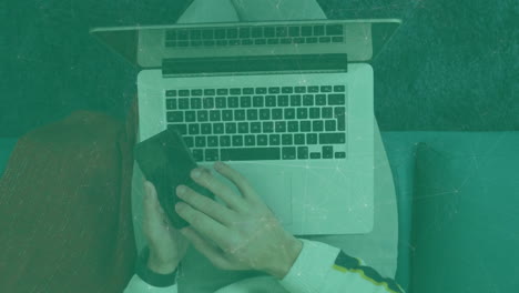 man using laptop and smartphone in living room, displaying animated finance charts and icons