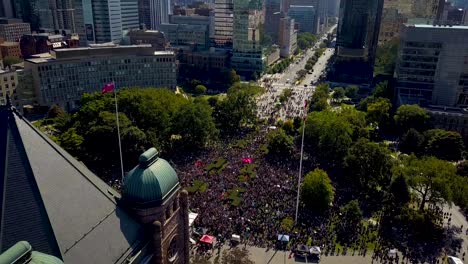 epic drone view of protesters in downtown toronto, aerial dolly out. big crowd of people at queens park is dwarfed by ontario parliament building in metaphor for democracy, wide daytime exterior in 4k