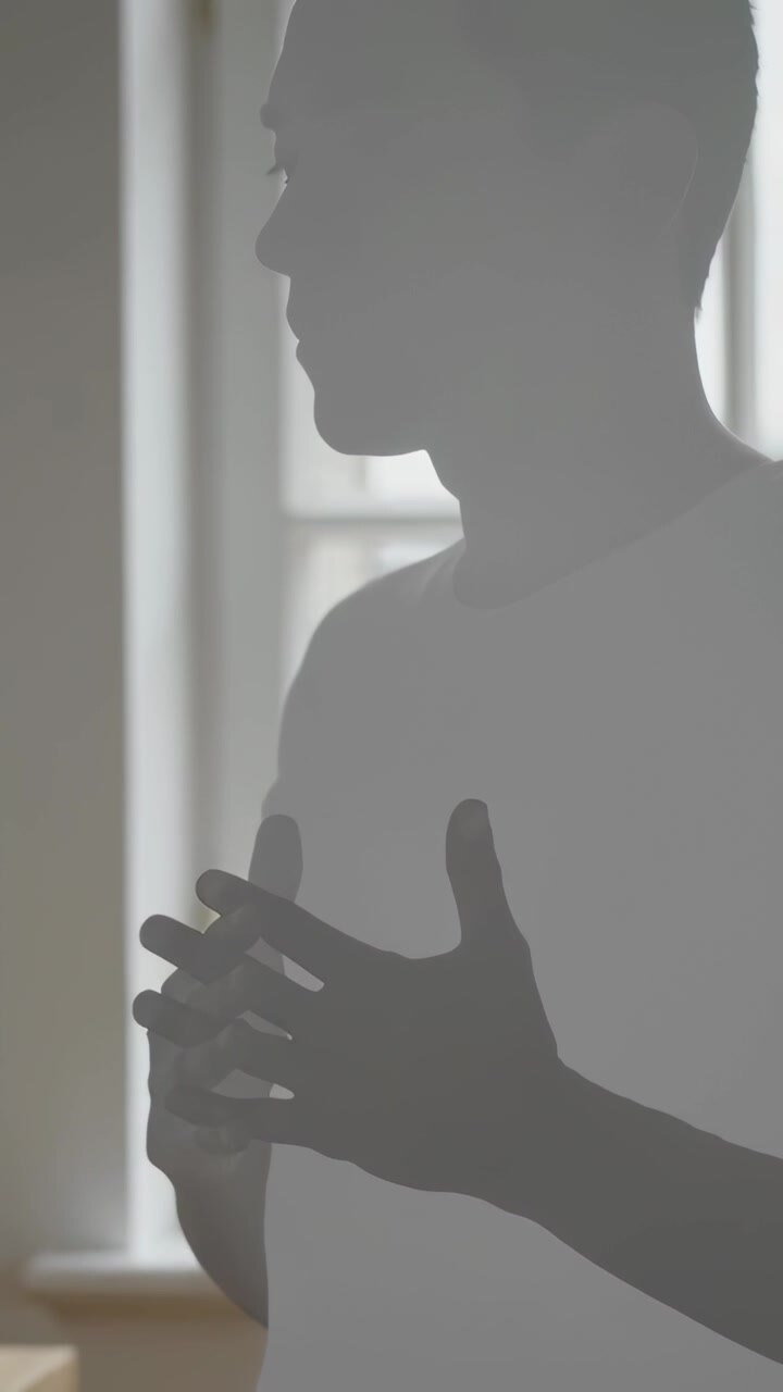 Vertical video: Responding to offscreen prompt, man gesturing near chest at window in light tee
