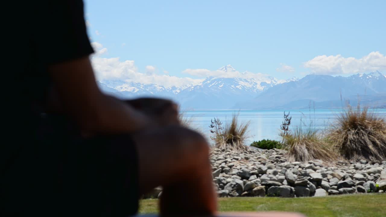 Young man sitting on outside bench drinking coffee while taking in breathtaking view of Mount Cook, New Zealand