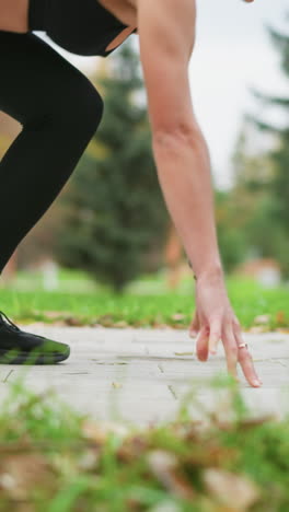 rotating view of athletic woman in black leggings getting ready to run outdoors, with hands on the ground, preparing for takeoff in park, focusing on her form against background of greenery