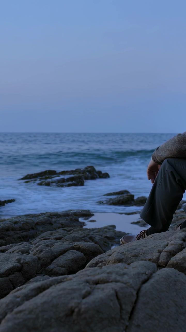 Vertical video: Sitting man in sweater pants gazing at waves, rocky tide pools at dusk, copy space