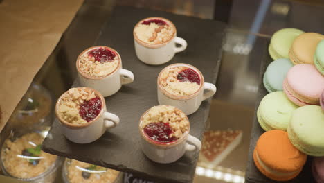 close up of delicious dessert cups filled with berry sauce and crumbles next to stacked colorful macarons inside store display, showcasing vibrant sweet treats under warm lighting in bakery