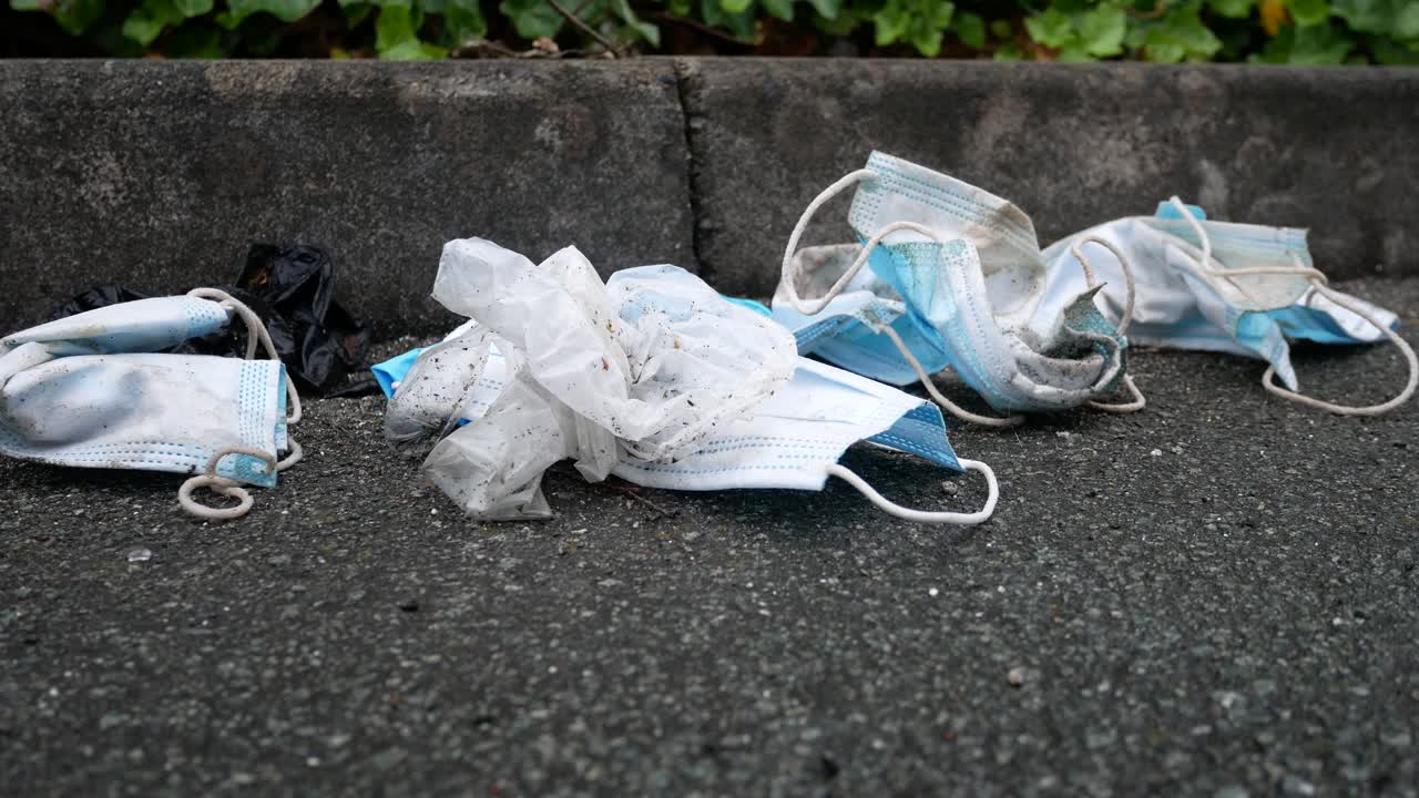 더럽게 버려진 사용된 코로나 바이러스 마스크는 거리 보도에서 바람에 쓰레기를 불고 있습니다.