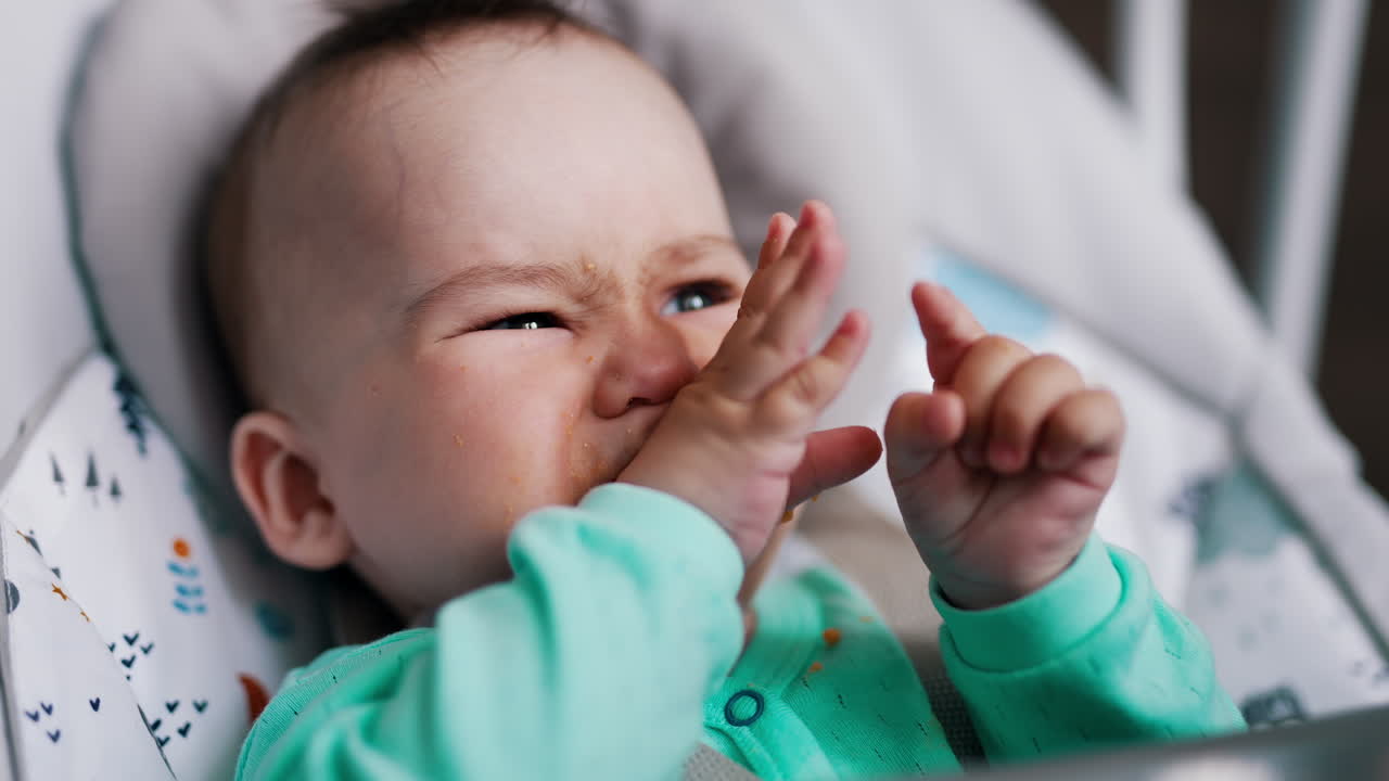 Mother's feeding her little baby from spoon. Cute kid taking food and wrinkles up and rubs his eyes. Close up.