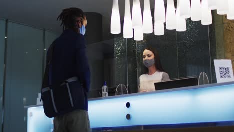 Mixed-race-man-and-female-receptionist-wearing-face-masks-at-hotel-reception-desk