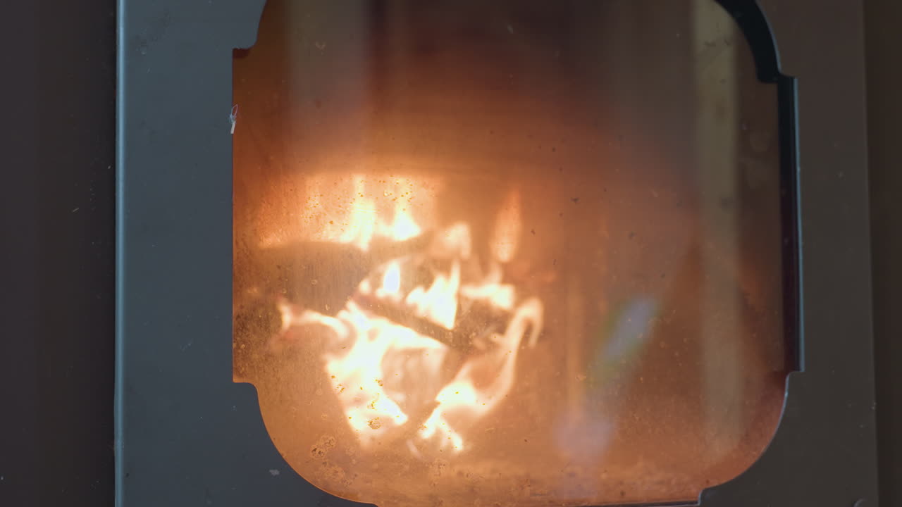 Firewood burning intensely inside chimney viewed through slightly smudged glass door, flames flicker and illuminate enclosed space with soft orange glow, casting reflections and shadows on metal