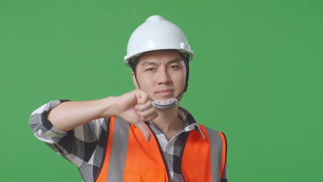 close up of asian male engineer with safety helmet showing thumbs down gesture and shaking his head while standing in the green screen background studio