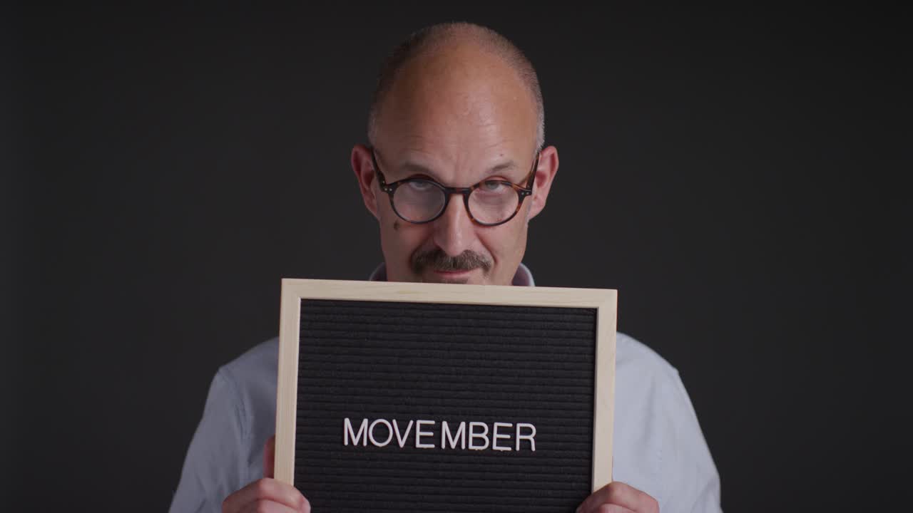 Studio Portrait Of Mature Man Holding Up Sign Reading Movember Promoting Awareness Of Men's Health And Cancer