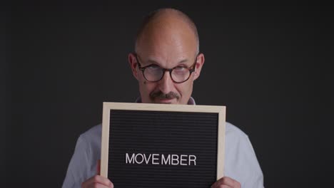 Studioporträt-Eines-Reifen-Mannes,-Der-Ein-Schild-Mit-Der-Aufschrift-„Movember“-Hochhält-Und-Das-Bewusstsein-Für-Männergesundheit-Und-Krebs-Fördert