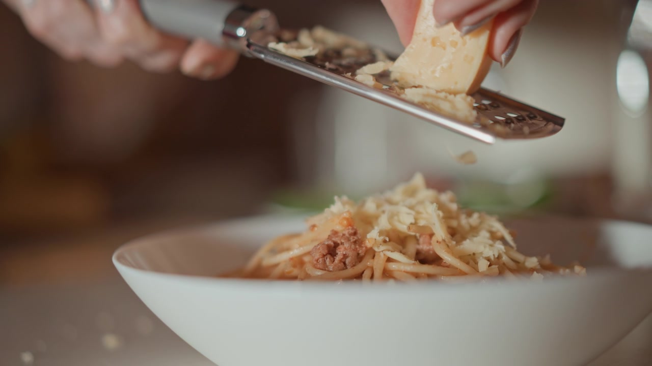 Scena di cucina ravvicinata dal basso che mostra una mano che grattugia formaggio giallo pallido direttamente sopra i noodles conditi con salsa in una ciotola di ceramica bianca, con scaglie che cadono, minuscole particelle sospese nell'aria e un pasto caldo e invitante
