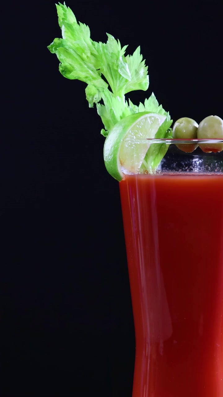 Um coquetel Bloody Mary com aipo, limão e azeitonas contra um fundo escuro. Câmera estática, cores vibrantes e apresentação elegante.