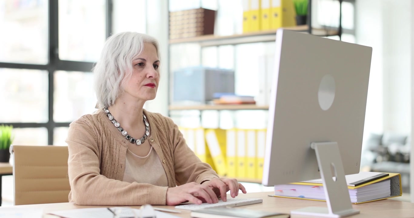 Senior woman at desk with computer actively types on keyboard