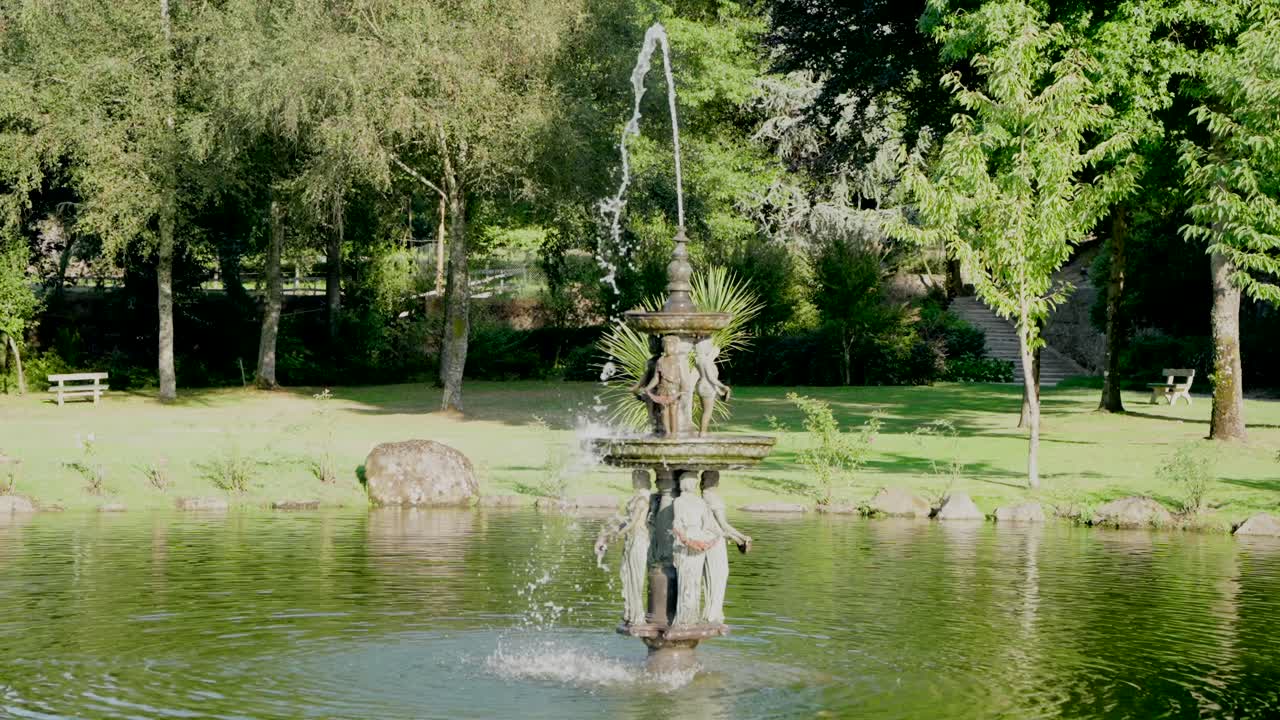 decorative fountain in the middle of a pond, surrounded by trees in a peaceful park setting