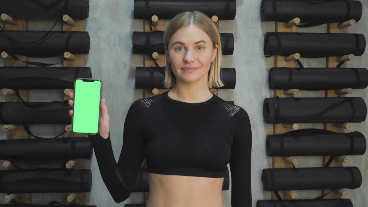 una joven atlética sostiene un teléfono inteligente en la mano, muestra una pantalla chromakey verde, está en el gimnasio