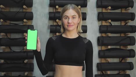 a young athletic woman holds a smartphone in her hand, shows a green chromakey screen, is in the gym