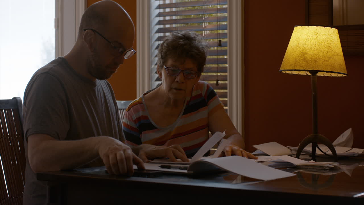 A middle-aged man, and senior woman sit together at a dining table in a warmly lit home, sorting through bills. Son is focused on reviewing receipts, Onlooking mother has difficulty seeing.
