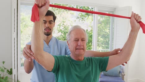 Biracial-male-physiotherapist-with-caucasian-senior-man-holding-resistance-band-while-exercising