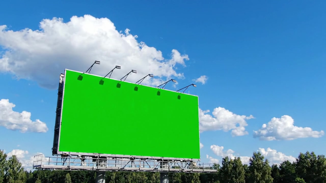 En tom grøn billboard, der står under den lyse blå himmel, omgivet af frodige grønne træer og bløde hvide skyer, perfekt til reklameplads og kampagner