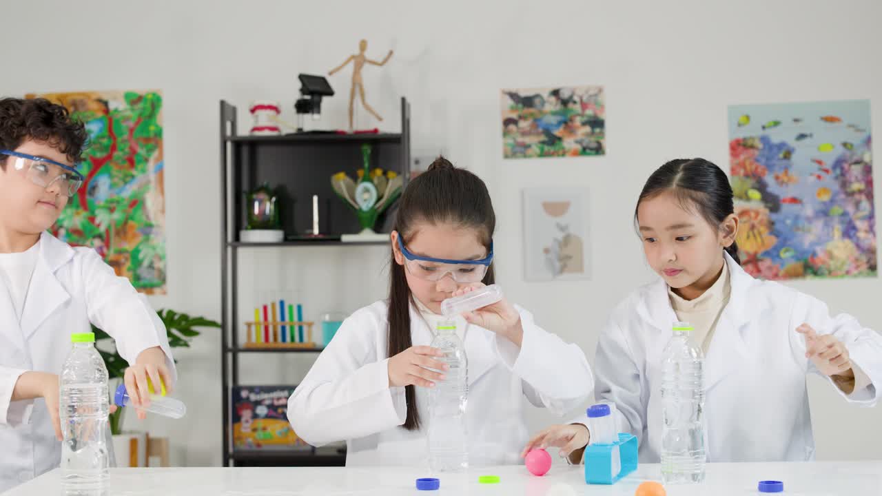 白衣を身につけた3人のアジア系の子どもたちが、瓶とボールを使って物理実験を行っています。