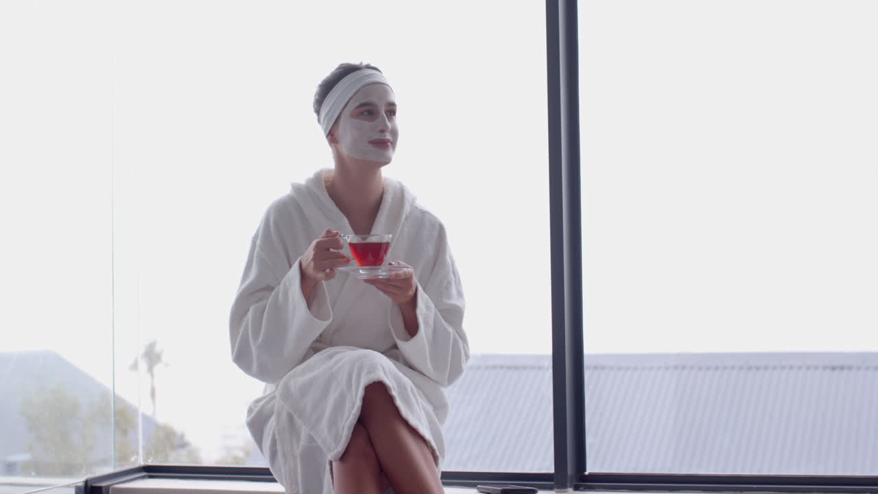Wearing face mask, woman in bathrobe drinking tea by window, at home, copy space