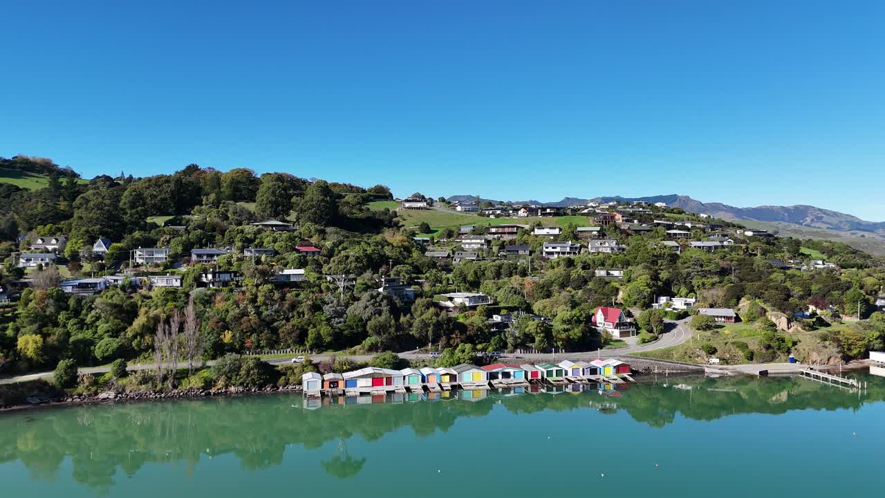Drone footage captures Akaroa's colorful waterfront houses and lush greenery under clear skies, reflecting in calm waters