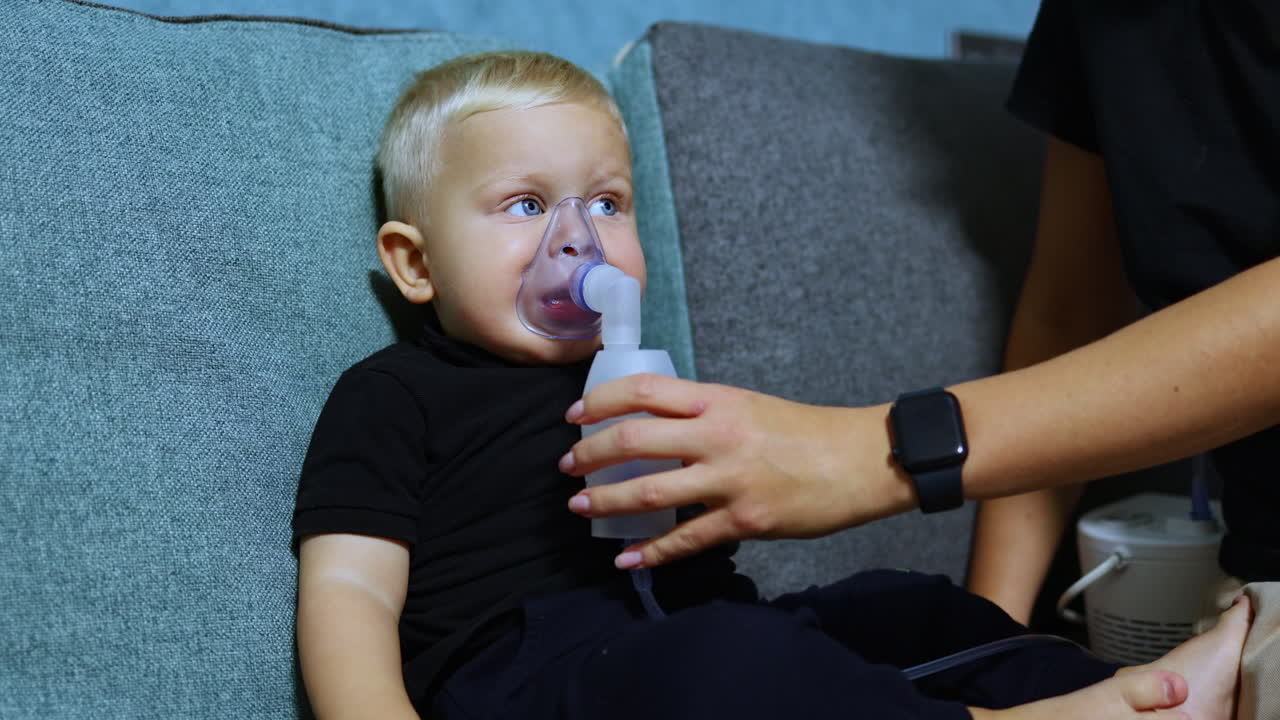 Malý chlapec sedí na gauči a inhaluje pomocí inhalátoru. Terapeutická inhalace pro děti. Inhalační maska pro děti.