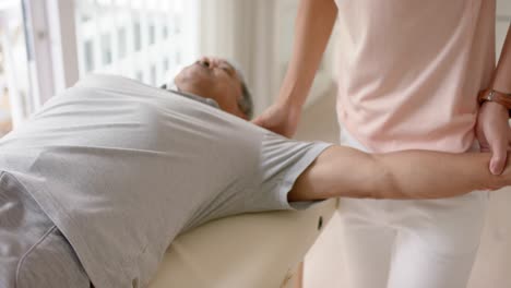 diverse male physiotherapist advising and senior male patient exercising on couch, in slow motion