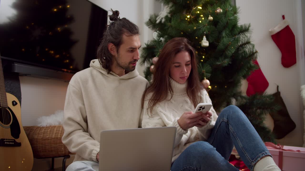 un chico y una chica morenas serios y reflexivos con suéteres blancos se sientan en el suelo y trabajan en una computadora portátil y teléfono cerca del árbol de año nuevo en una habitación acogedora en una noche de invierno