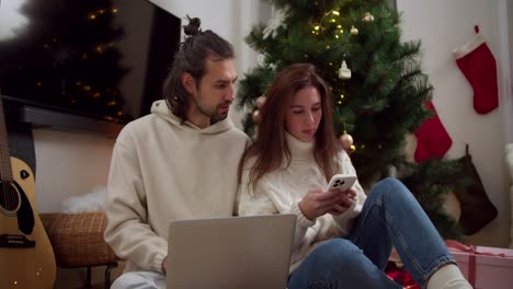 Serious-and-thoughtful-brunette-guy-and-girl-in-white-sweaters-sit-on-the-floor-and-work-on-a-laptop-and-phone-near-the-New-Year-tree-in-a-cozy-room-in-a-winter-evening