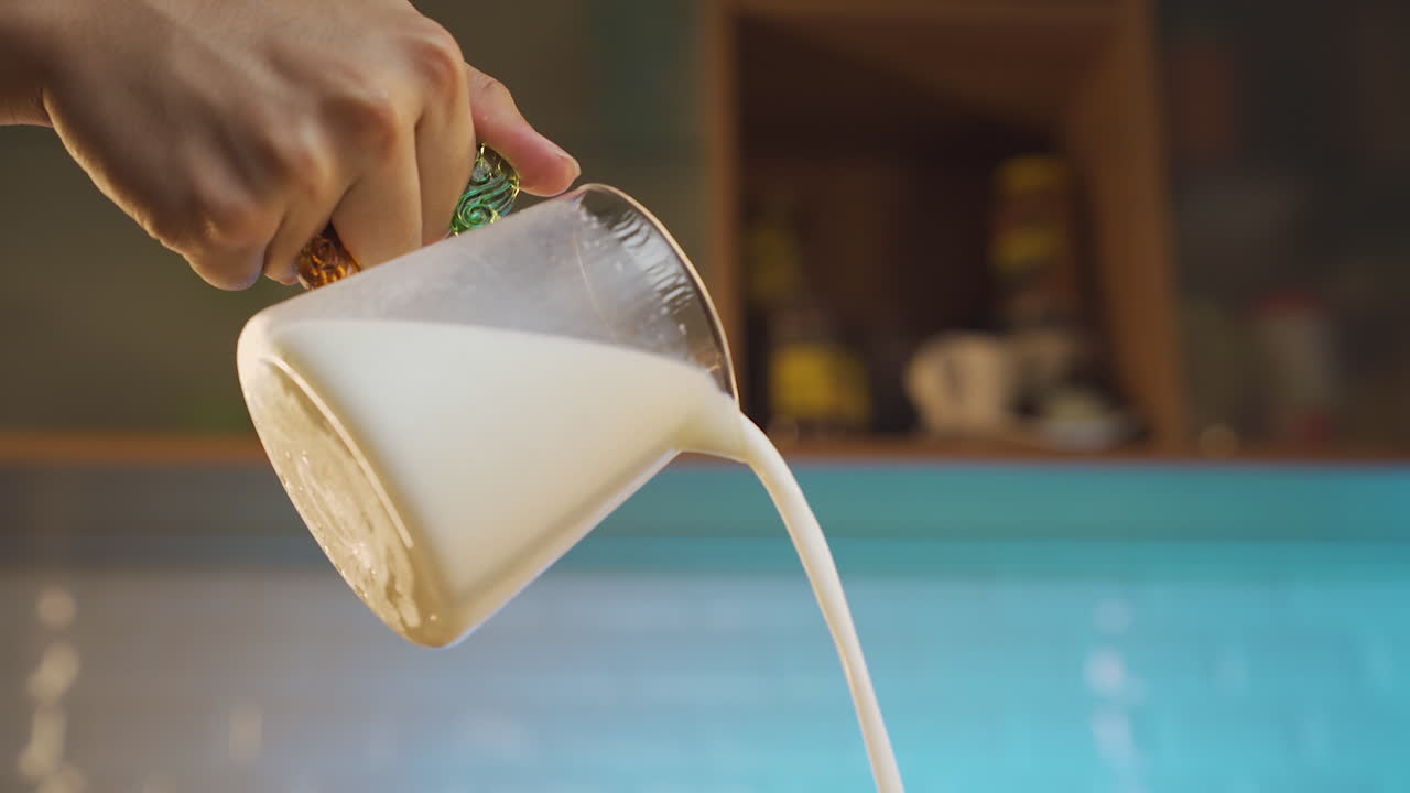 a person is pouring milk from a glass container with blue ambient kitchen in the background.