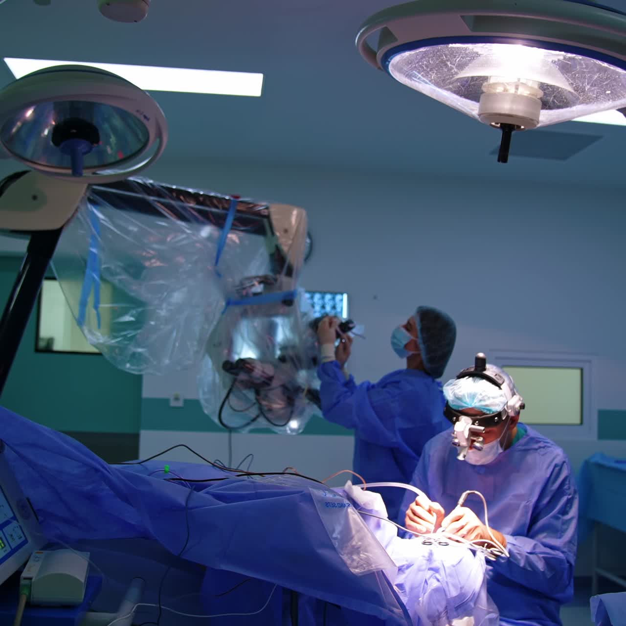 Surgeon wearing surgical loupes works at operation using tools. Nurse at backdrop prepares the microscope for usage by the doctor