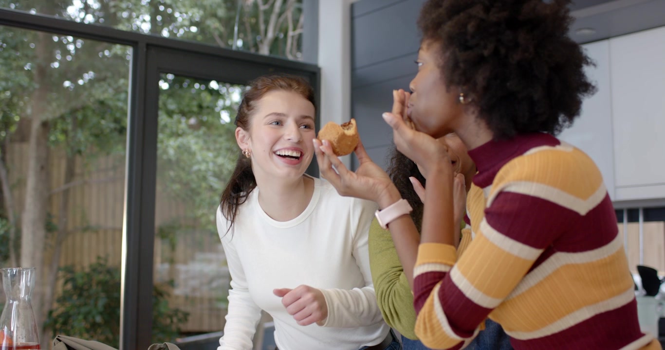 Three women pals watching woman in striped top picking muffin testing recipe in kitchen, copy space