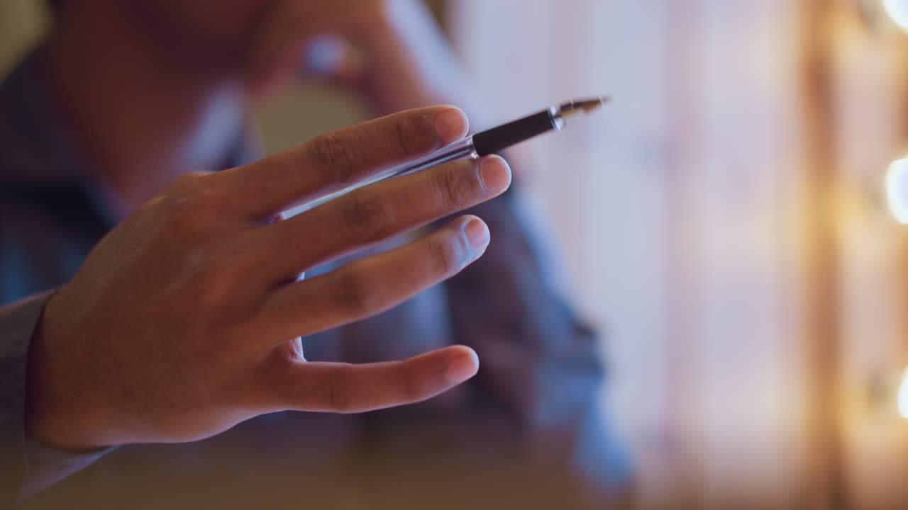 Close-up of man holding pen while sitting thoughtfully near window with soft glowing lights in background, suggesting contemplation, planning, or creative brainstorming in cozy ambient setting
