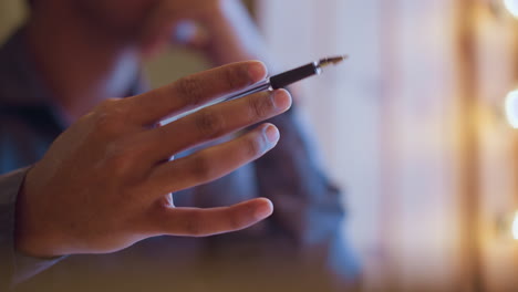 close-up of man holding pen while sitting thoughtfully near window with soft glowing lights in background, suggesting contemplation, planning, or creative brainstorming in cozy ambient setting