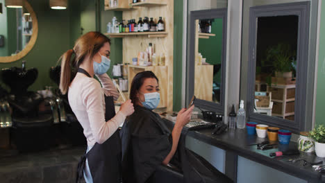 female hairdresser and female customer wearing face masks looking at smartphone at hair salon