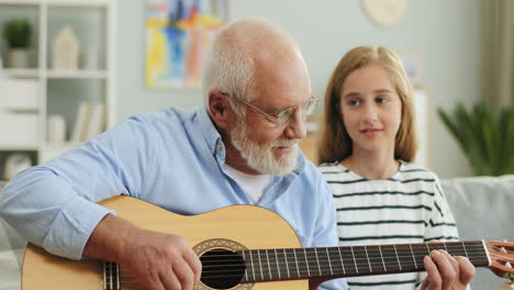 Portrait-Shot-Of-The-Old-Grandfather-In-Glasses-Playing-The-Guitar-And-His-Granddaughter-Listening-To-Him-At-Home