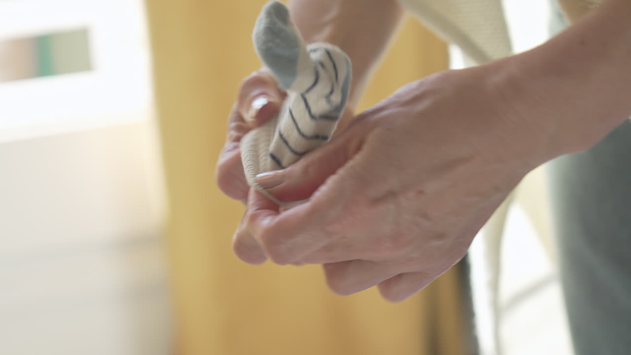 madre recoge calcetines de bebé a rayas, vistiendo al bebé, en un dormitorio blanco iluminado por el sol