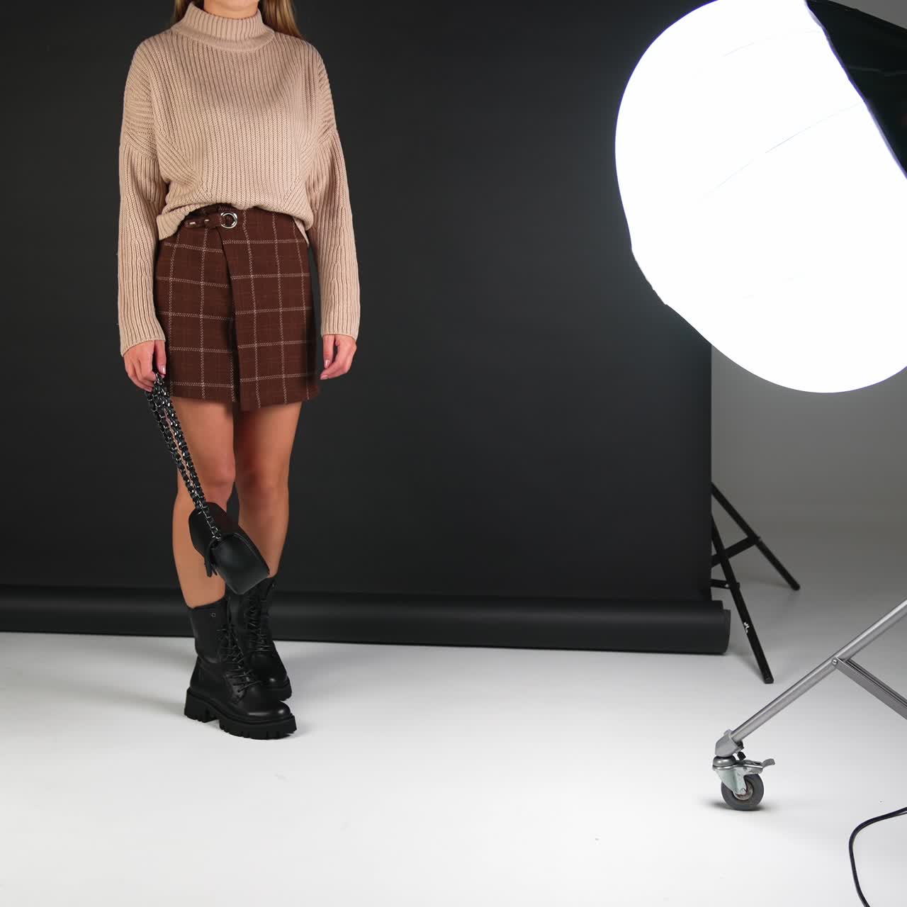 Young female model wearing sweater, mini skirt and high black boots poses in studio. Lady demonstrating the shoes and bag