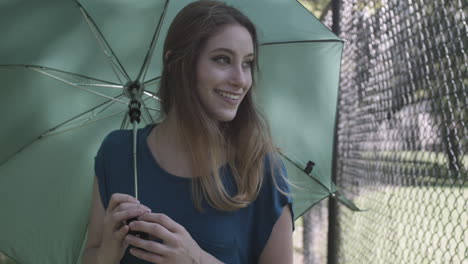 Young-millennial-woman-walks-next-to-a-chain-link-fence-with-a-green-umbrella-on-a-sunny-day