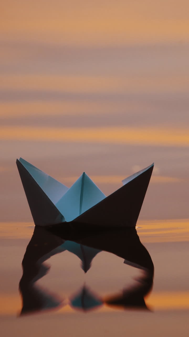 Origami boat on water surface in the evening. White paper boat swims with its shadow reflection in water. Close-up. Vertical video