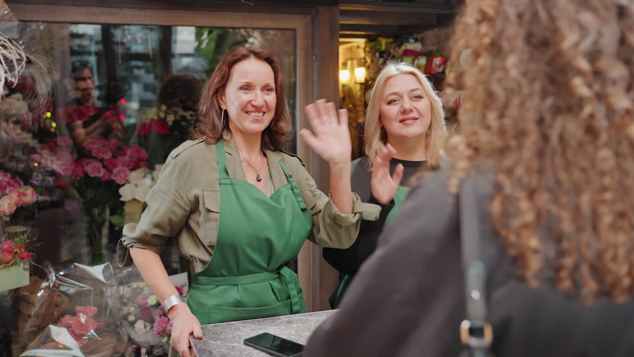 Duo di fioristi dietro il bancone sorridono e salutano una cliente che ritorna, grembiuli verdi, abbondanti esposizioni floreali, rose e tulipani, vetrina rustica, calda atmosfera da mercato di quartiere, amichevole